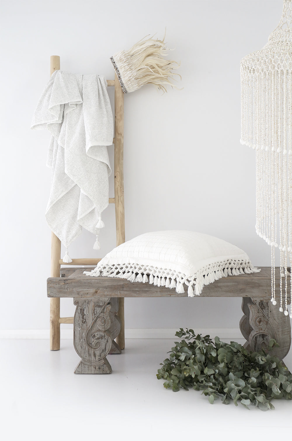 A beautifully styled space featuring a rustic wooden bench with a white textured cushion, a natural wooden ladder draped with soft tassel-edged towels, and a macramé pendant light. A neutral-toned, coastal-inspired setting with organic textures