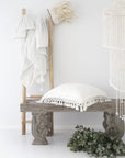 A beautifully styled space featuring a rustic wooden bench with a white textured cushion, a natural wooden ladder draped with soft tassel-edged towels, and a macramé pendant light. A neutral-toned, coastal-inspired setting with organic textures