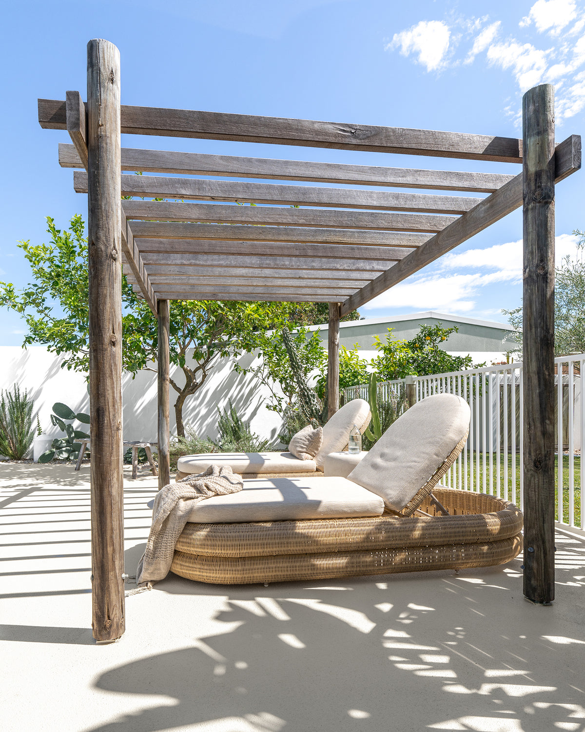 KULALA Living Sage Rectangle Linen Cushion and Cairo Linen Throw styled on natural woven outdoor sun loungers beneath a rustic timber pergola in a sunlit garden setting.