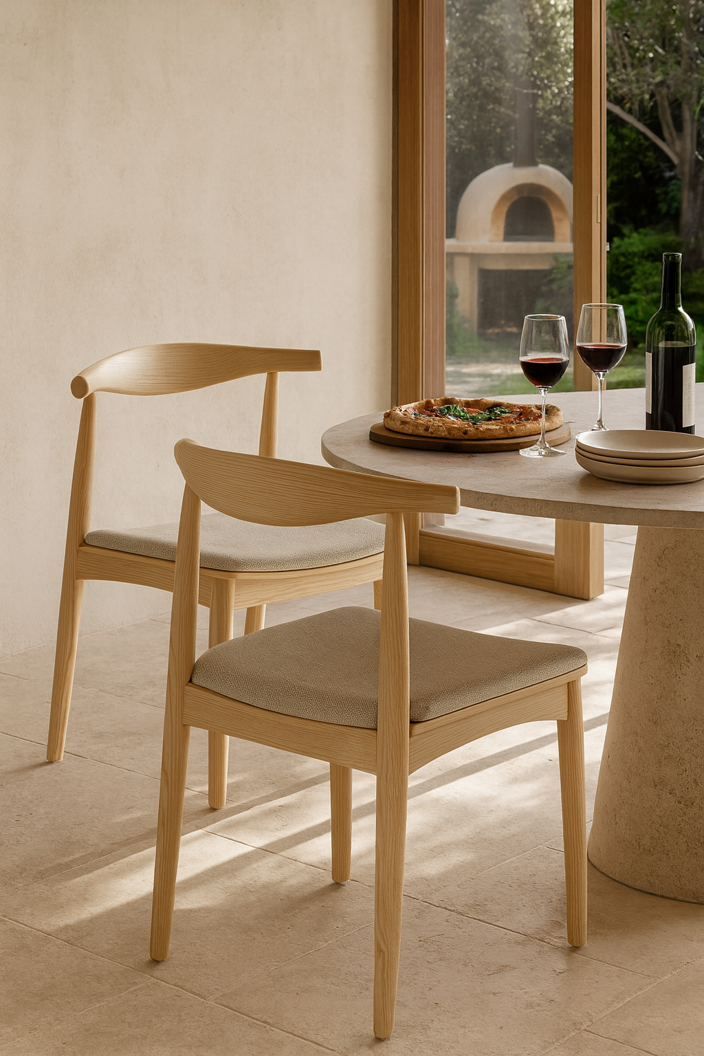 Pair of Scandinavian-style oak dining chairs with neutral upholstered seats styled beside a round stone table, served with pizza and red wine in a sunlit setting with garden views