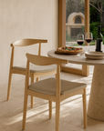 Pair of Scandinavian-style oak dining chairs with neutral upholstered seats styled beside a round stone table, served with pizza and red wine in a sunlit setting with garden views
