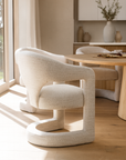 Curved sculptural dining chair upholstered in textured cream fabric, styled in a sunlit dining room with light timber floors and round oak table