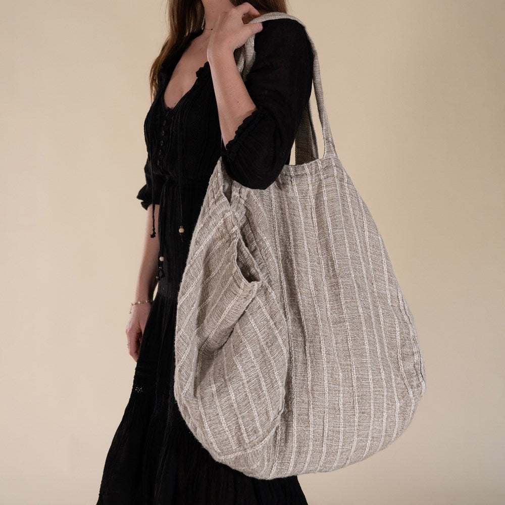 A woman in a black bohemian dress carrying a generously sized striped linen market bag with a relaxed, slouchy design and exterior pocket. The neutral-toned fabric and soft texture create a timeless, effortless style