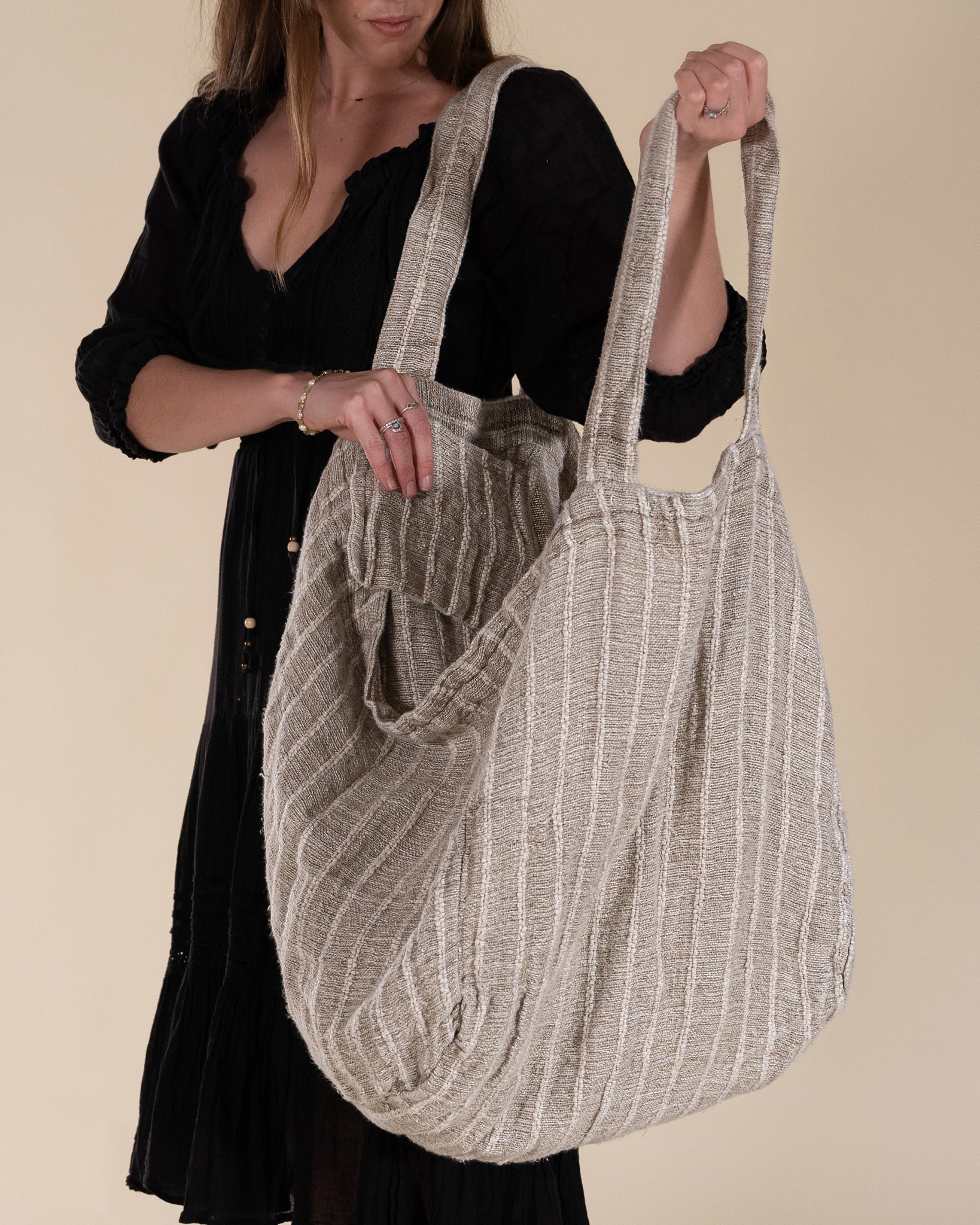 A woman in a black bohemian dress carrying a generously sized striped linen market bag with a relaxed, slouchy design and exterior pocket. The neutral-toned fabric and soft texture create a timeless, effortless style