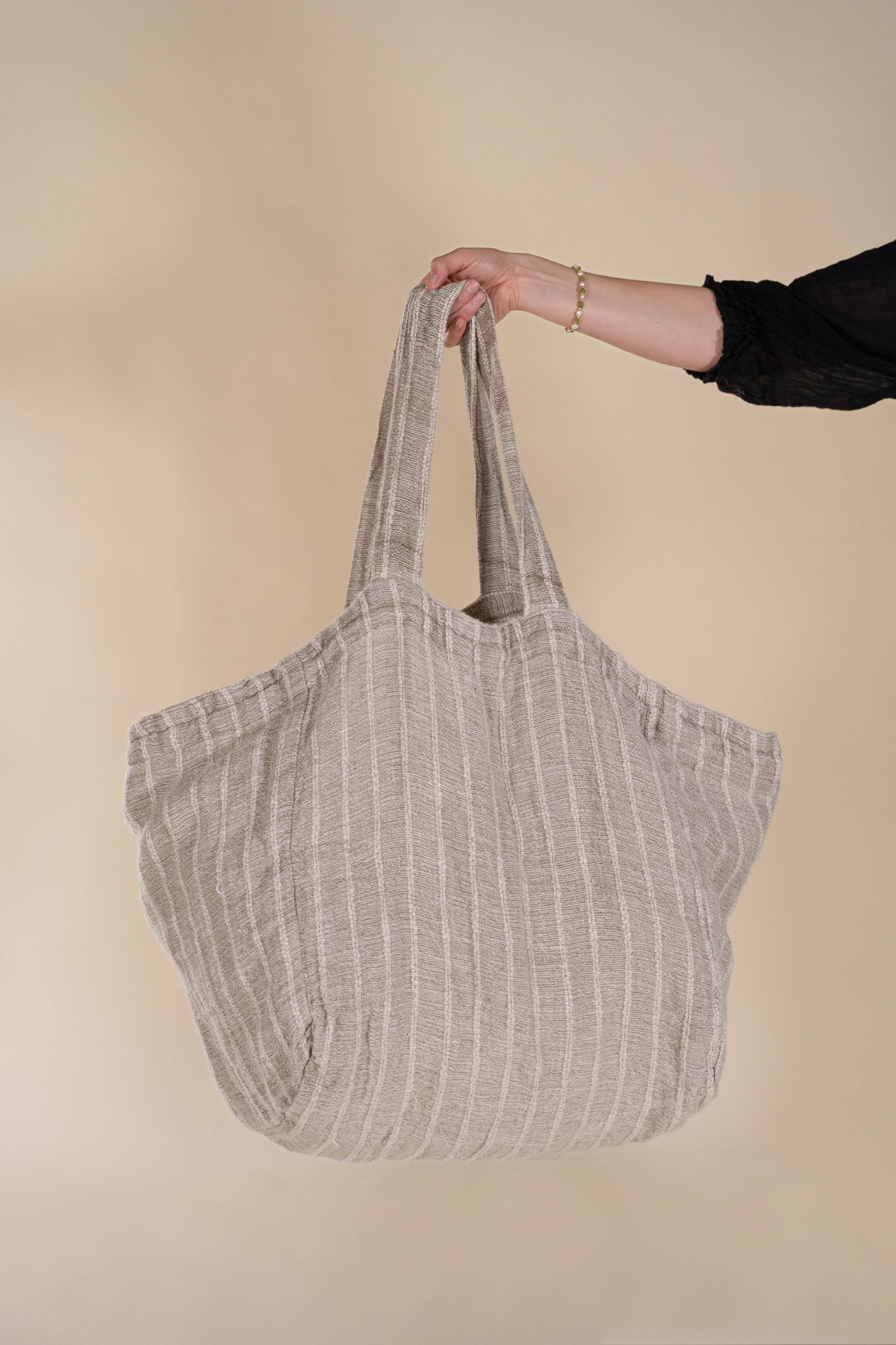 A woman in a black bohemian dress carrying a generously sized striped linen market bag with a relaxed, slouchy design and exterior pocket. The neutral-toned fabric and soft texture create a timeless, effortless style