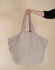 A woman in a black bohemian dress carrying a generously sized striped linen market bag with a relaxed, slouchy design and exterior pocket. The neutral-toned fabric and soft texture create a timeless, effortless style