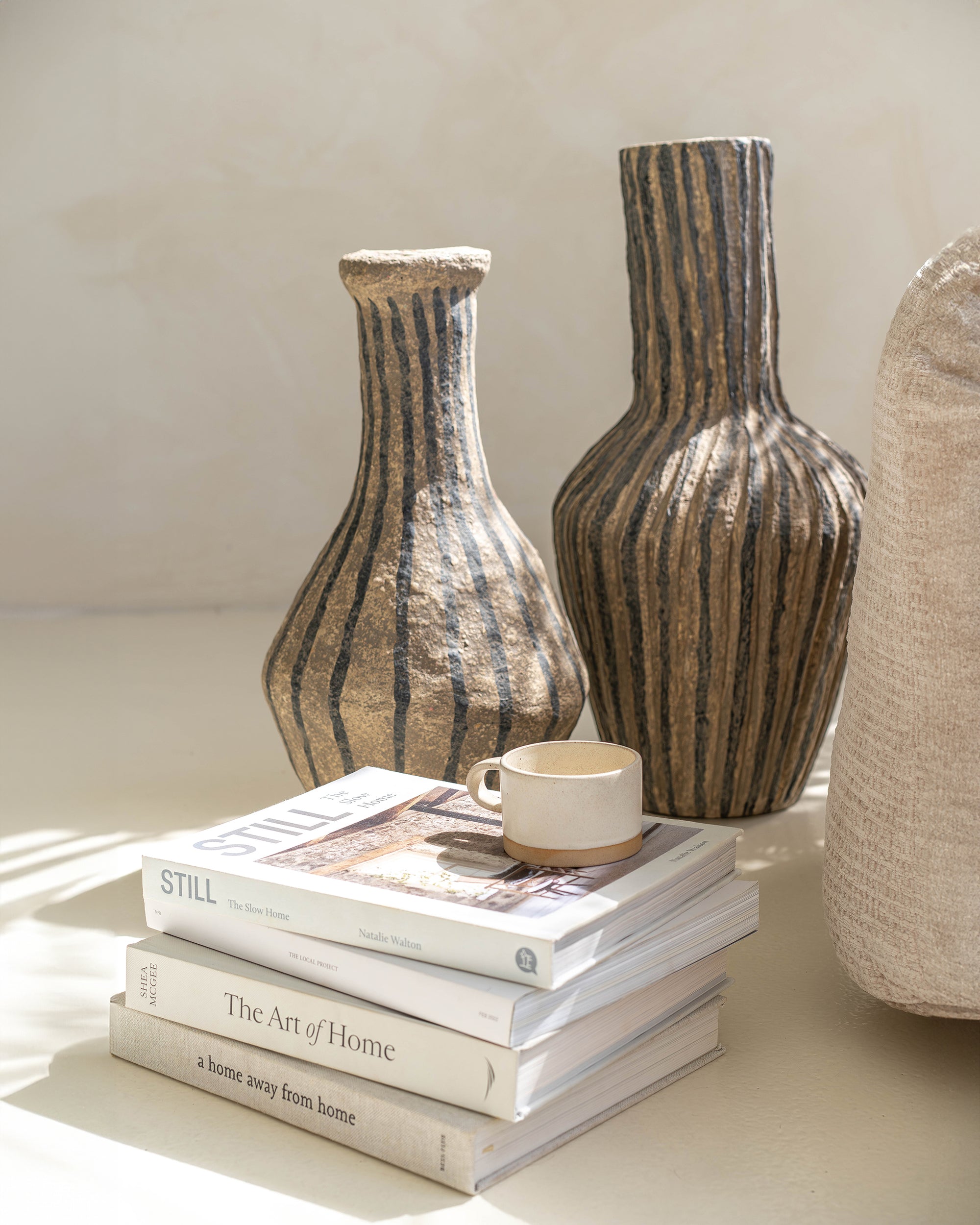 A beautifully styled vignette featuring two hand-sculpted papier mache vases with textured black stripes, stacked interior design books, and a minimalist ceramic cup. Soft natural light enhances the earthy tones, creating a warm and inviting aesthetic