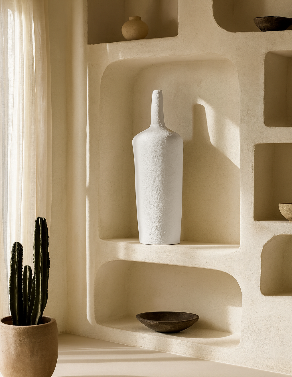 Tall white ceramic vase with a slender neck displayed on a sculptural built-in shelf, styled with earthy bowls and natural light.