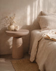 Tarran side table in nomad dune with sculptural pedestal form styled beside a bed in a soft sunlit bedroom setting.