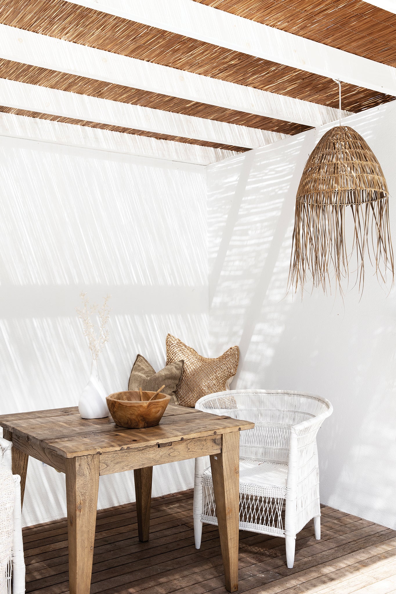 Natural warm brown teak carved bowl styled on a rustic timber outdoor dining table with white wicker chair, woven pendant and soft coastal light.