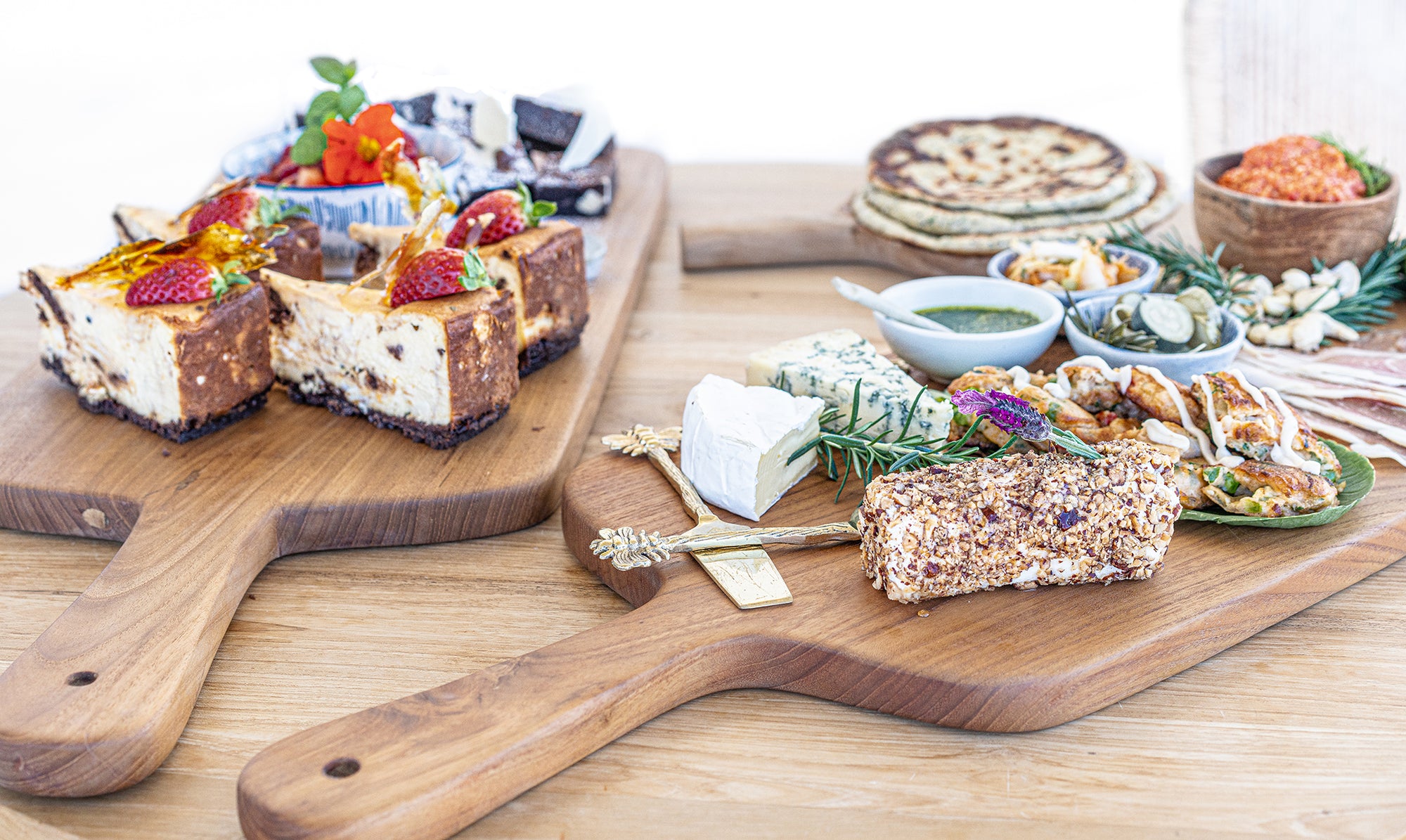 Teak carved chopping boards styled for entertaining with cheeses, desserts and small dishes arranged on a timber table in a bright setting.
