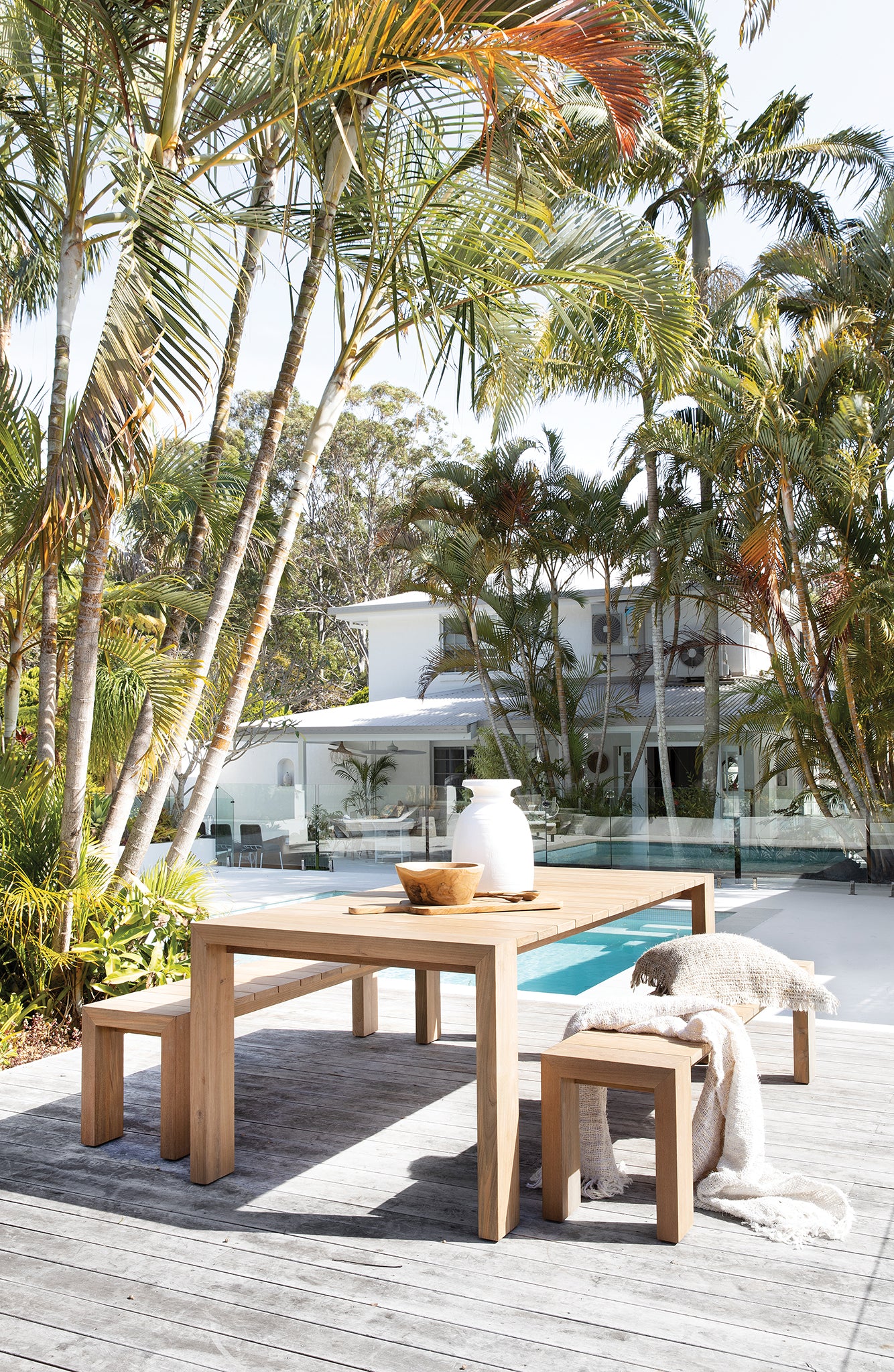 Teak carved chopping board styled on a solid timber outdoor dining table beside a pool, surrounded by lush palm trees and a relaxed coastal setting.