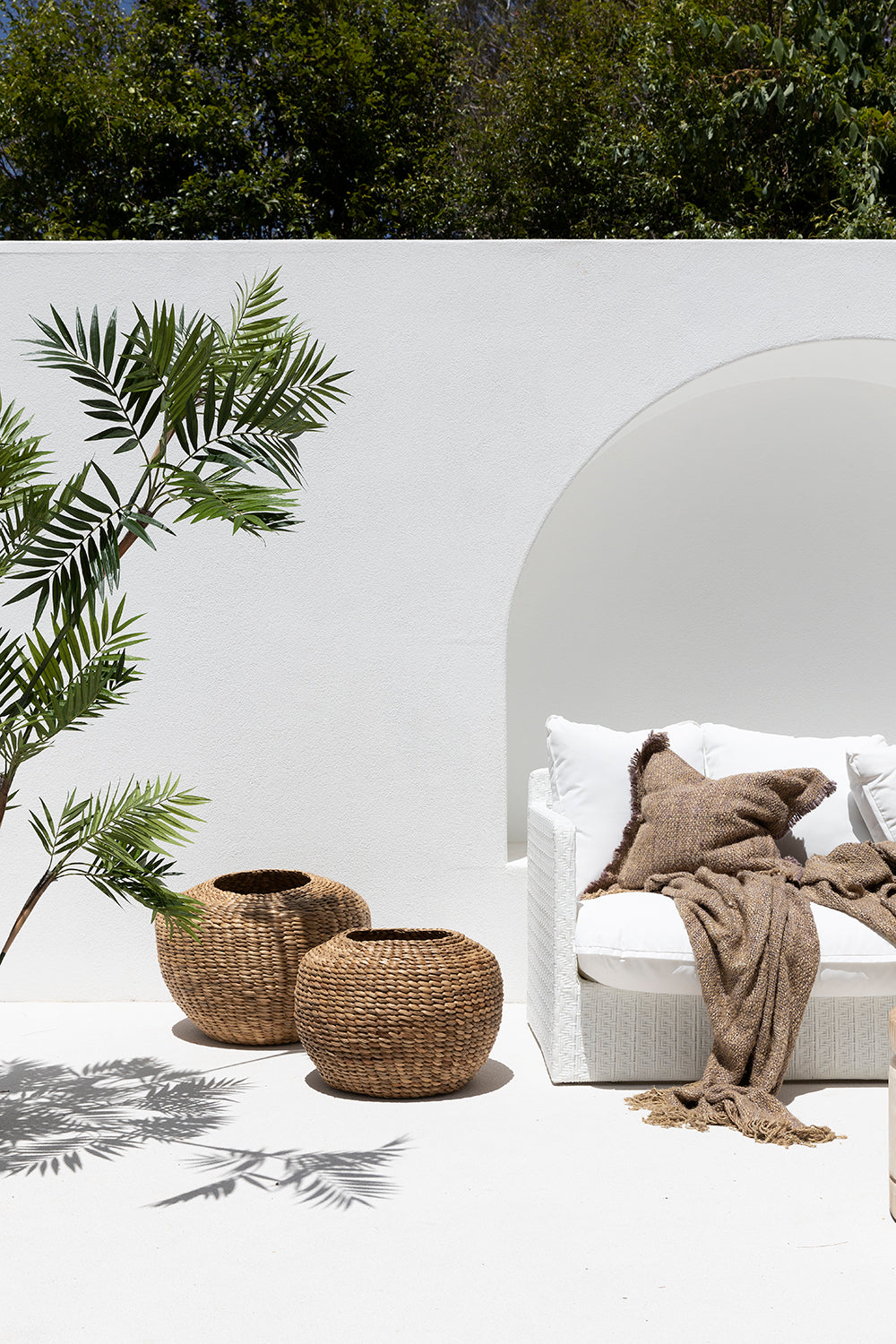 The Kulala Living Teak Drum Coffee Table styled in a bright outdoor setting. Crafted from natural teak branches, this round coffee table sits between two white lounge chairs with textured cushions, complemented by lush greenery and earthy decor for a serene alfresco retreat