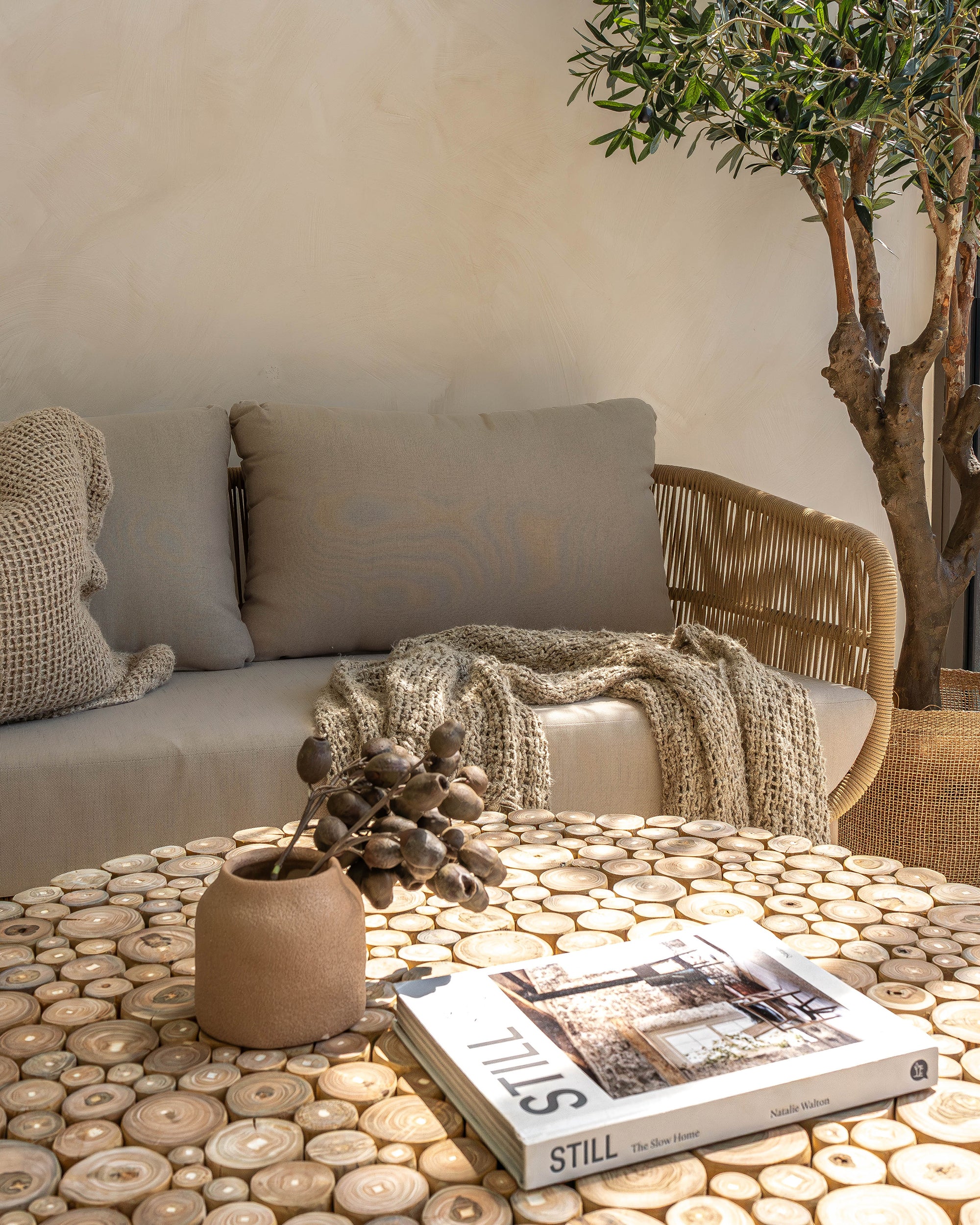 A close-up of the Kulala Living Teak Drum Coffee Table, showcasing its natural teak branch surface. Styled with a ceramic vase, dried botanicals, and the book Still: The Slow Home, the table sits in front of a woven lounge adorned with textured cushions and a knitted throw, creating a cosy and earthy living space