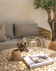 A close-up of the Kulala Living Teak Drum Coffee Table, showcasing its natural teak branch surface. Styled with a ceramic vase, dried botanicals, and the book Still: The Slow Home, the table sits in front of a woven lounge adorned with textured cushions and a knitted throw, creating a cosy and earthy living space