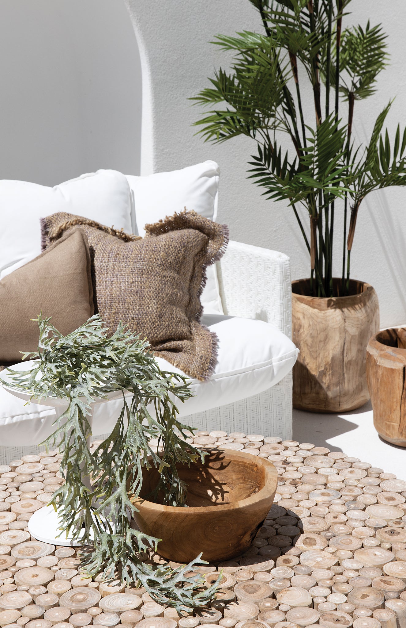 The Kulala Living Teak Drum Coffee Table styled in a bright outdoor setting. Crafted from natural teak branches, this round coffee table sits between two white lounge chairs with textured cushions, complemented by lush greenery and earthy decor for a serene alfresco retreat
