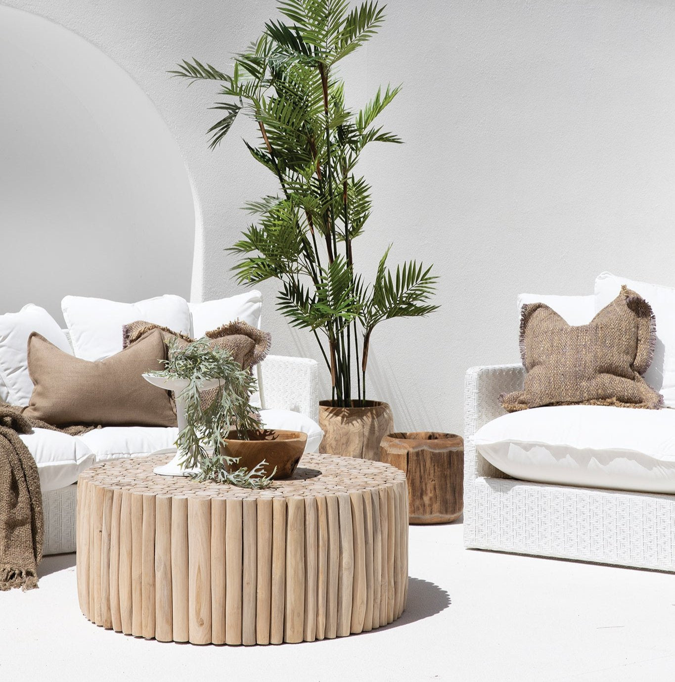 The Kulala Living Teak Drum Coffee Table styled in a bright outdoor setting. Crafted from natural teak branches, this round coffee table sits between two white lounge chairs with textured cushions, complemented by lush greenery and earthy decor for a serene alfresco retreat