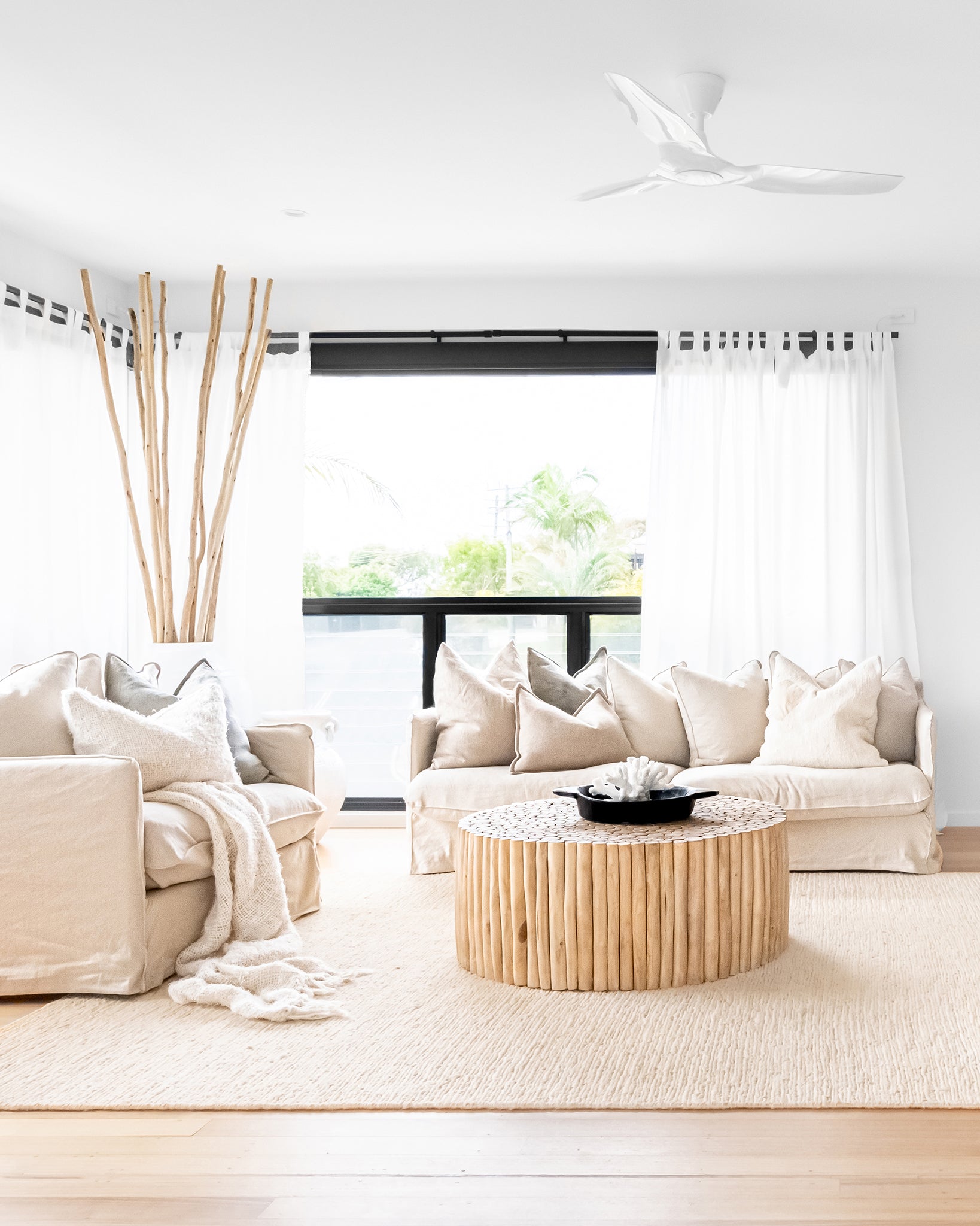 A bright and airy living room featuring the Kulala Living Teak Drum Coffee Table, styled with a decorative black bowl and coral accent. The space is furnished with plush neutral-toned sofas adorned with textured cushions and a soft throw, complemented by sheer white curtains and natural wooden decor for a serene, coastal-inspired aesthetic.