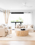 A bright and airy living room featuring the Kulala Living Teak Drum Coffee Table, styled with a decorative black bowl and coral accent. The space is furnished with plush neutral-toned sofas adorned with textured cushions and a soft throw, complemented by sheer white curtains and natural wooden decor for a serene, coastal-inspired aesthetic.