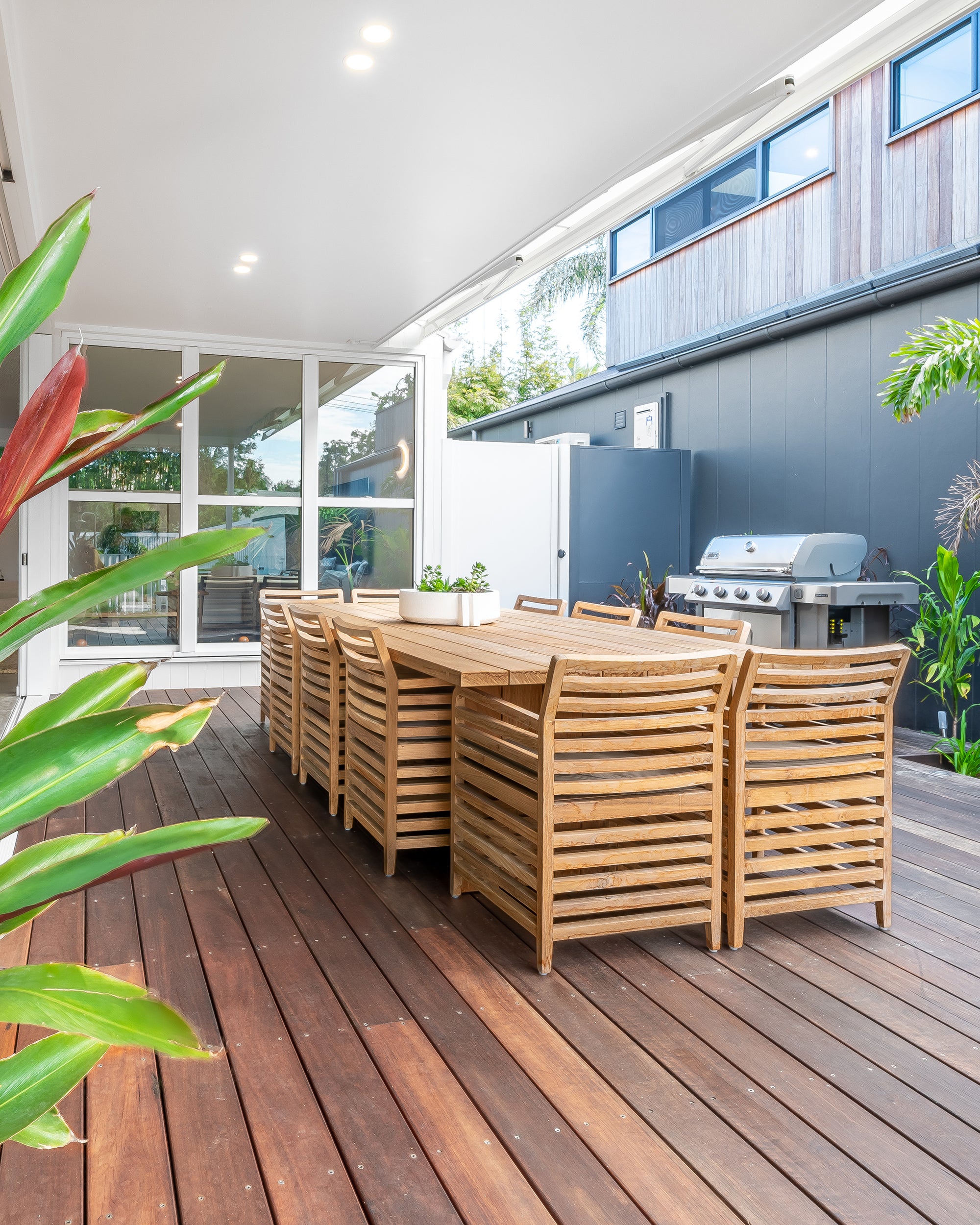 A spacious outdoor dining setting featuring a solid teak table and matching slatted dining chairs, designed for elegant alfresco entertaining on a timber deck