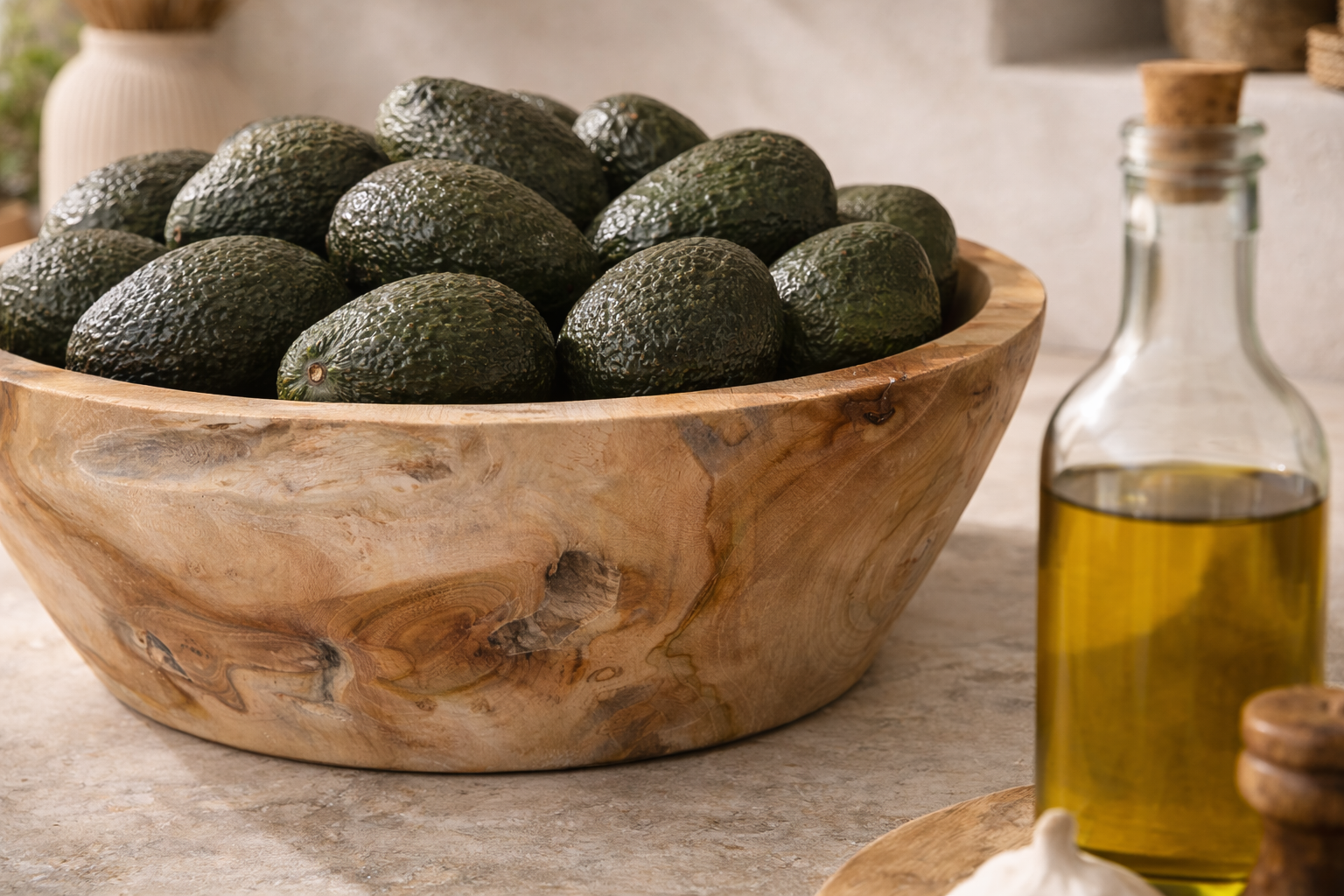 Large natural teak serving bowl filled with avocados, styled on a stone kitchen bench with neutral ceramics and olive oil in the background.