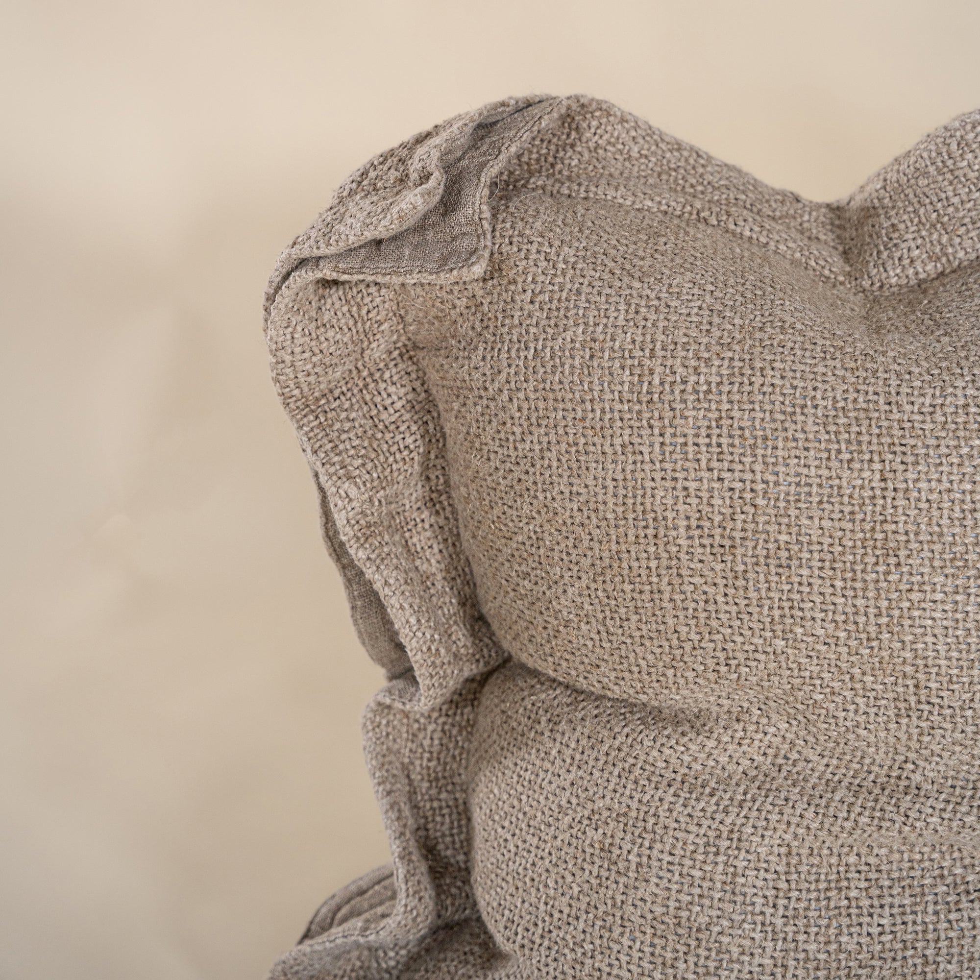 A textured linen cushion in a natural beige tone, featuring a relaxed, oversized design with raw-edge detailing, set against a neutral backdrop