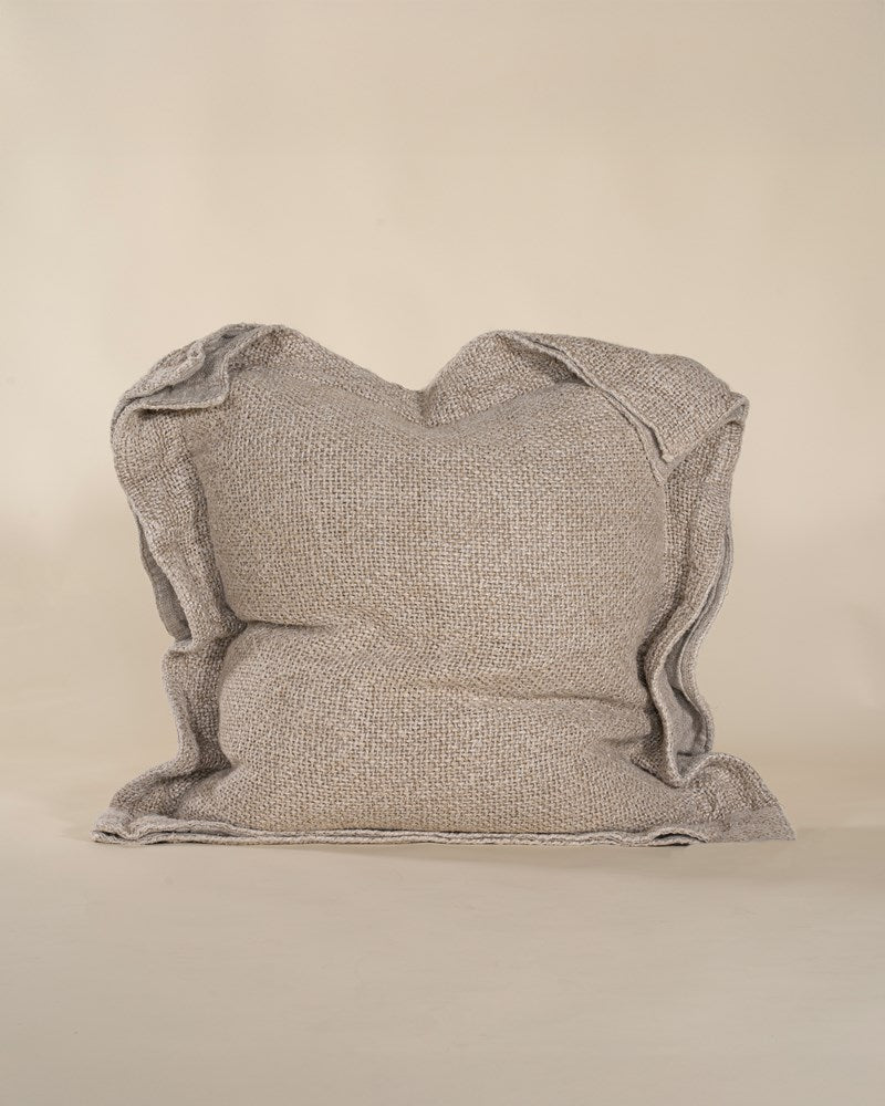 A textured linen cushion in a natural beige tone, featuring a relaxed, oversized design with raw-edge detailing, set against a neutral backdrop