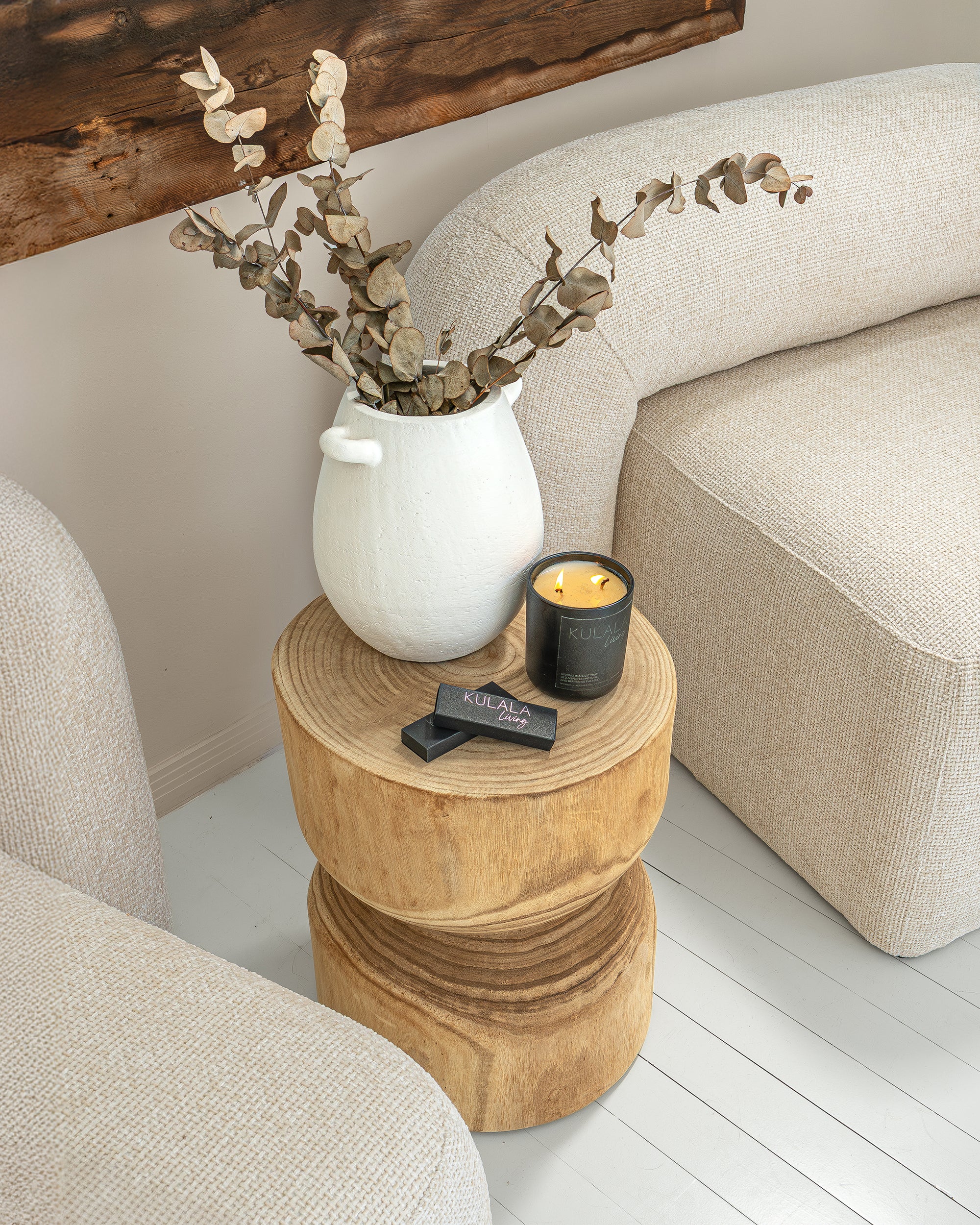 Close-up of a timber side table styled with a white ceramic vase, dried eucalyptus, KULALA Living black candle, and matches nestled between two boucle nash armchairs.