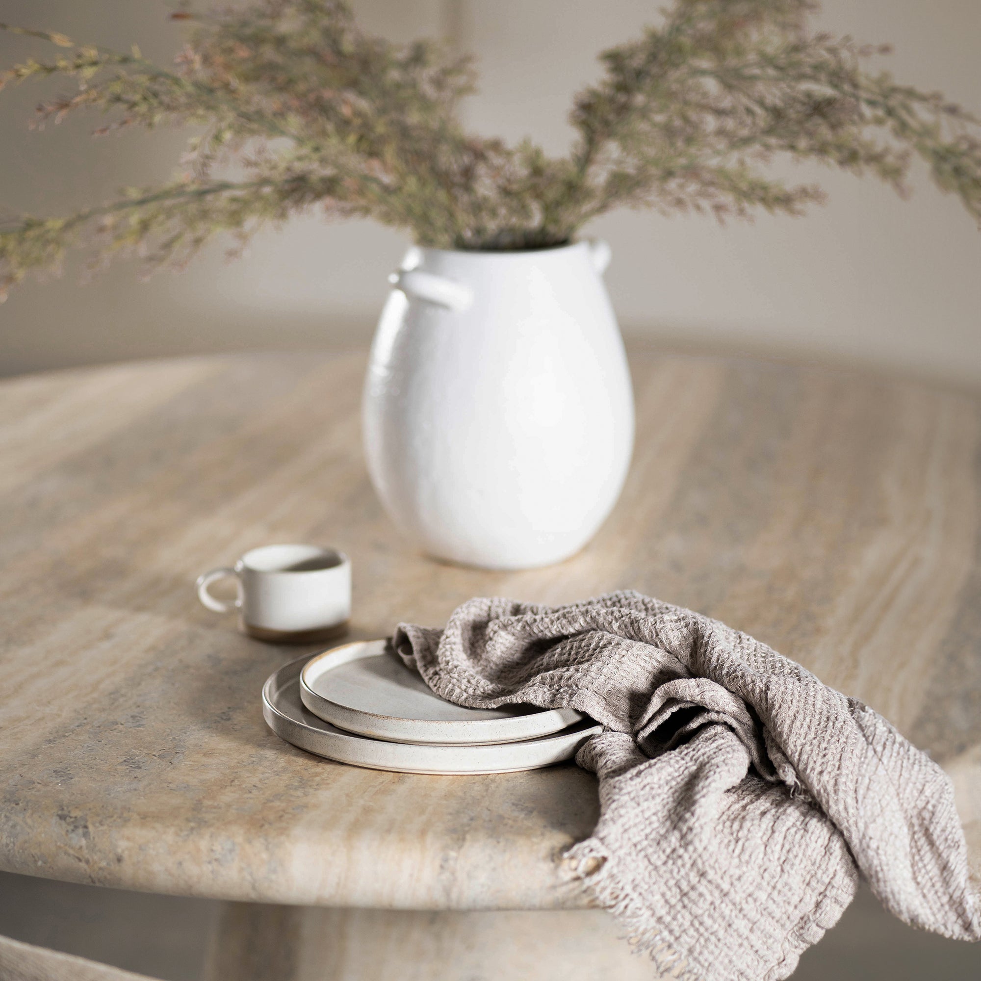 A beautifully styled dining space featuring a round travertine dining table, a neutral upholstered dining chair with timber accents, and a white ceramic vase with dried foliage. A ceramic cup, stacked plates, and a soft linen napkin add a touch of understated elegance