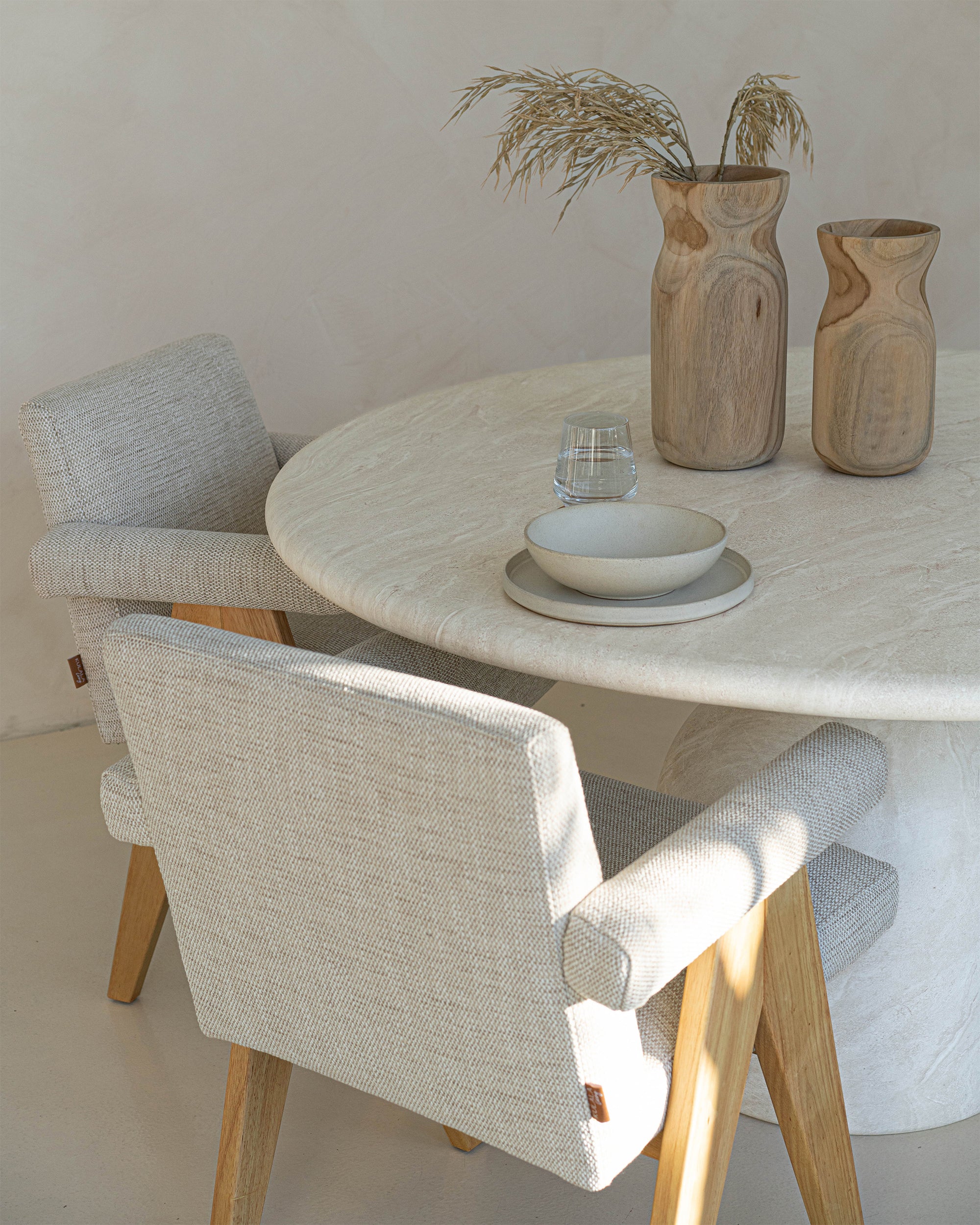 A serene dining space featuring a round travertine table with soft-textured upholstered chairs in a neutral palette. Wooden vases with dried foliage and minimalist ceramic tableware add organic warmth to the setting