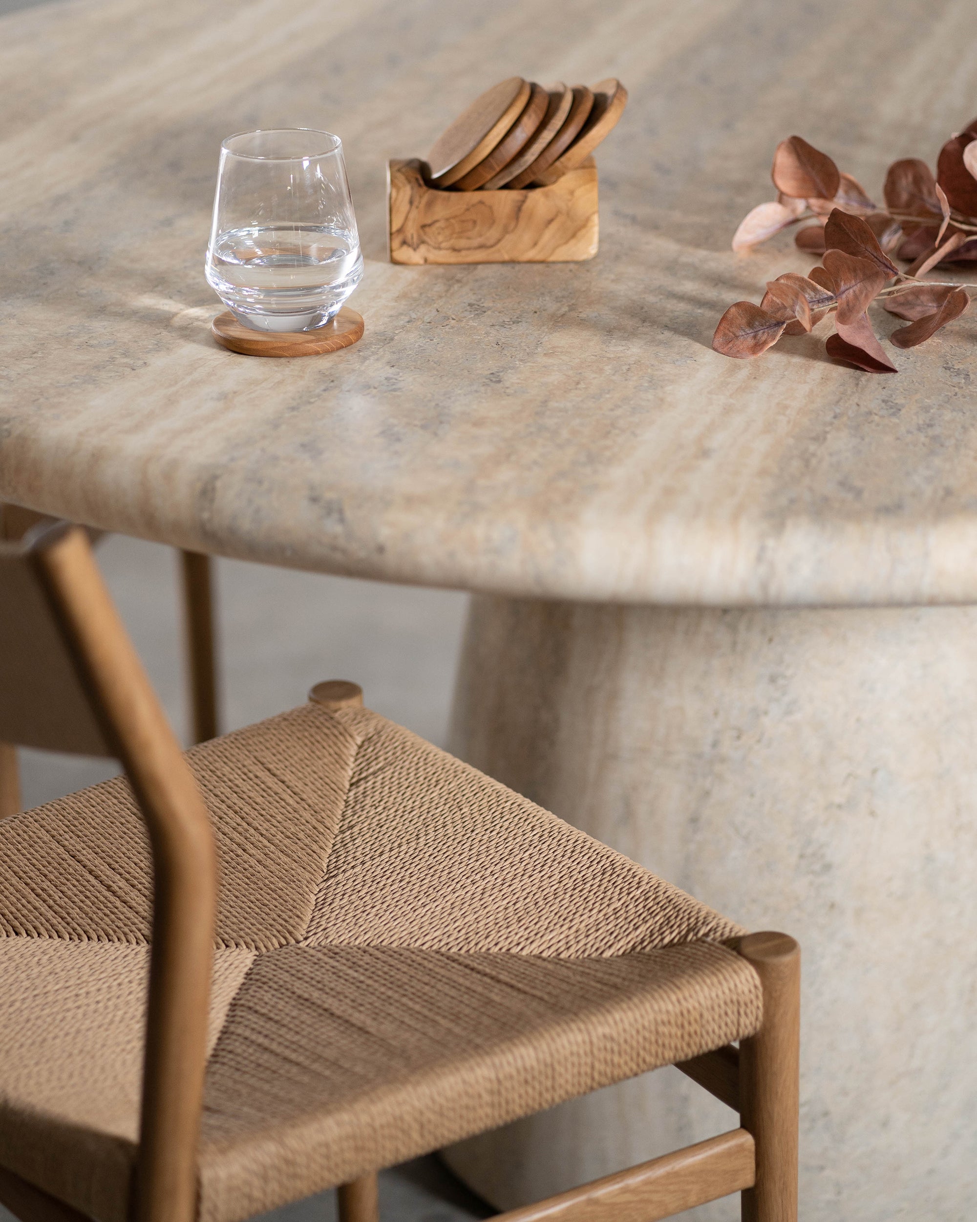 A beautifully styled travertine dining table featuring a wooden coaster set, a glass of water, and natural dried foliage, complemented by a rattan woven dining chair