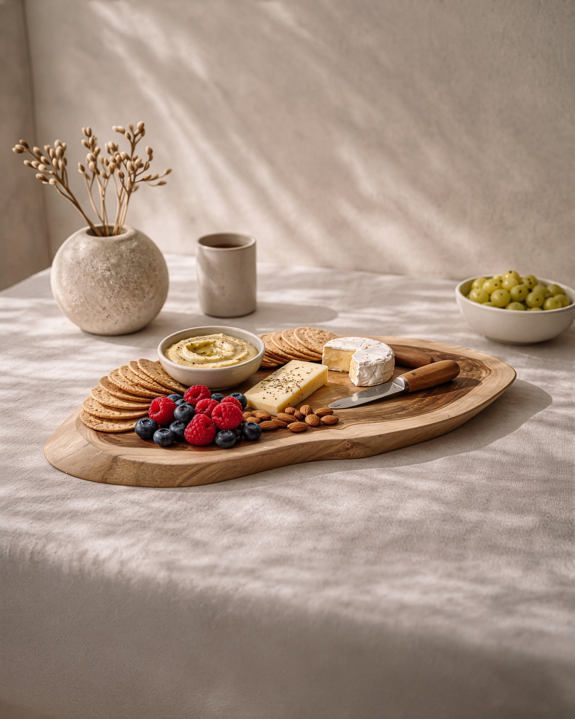 Vara cheese board in teak styled with cheese, grapes and crackers on a soft neutral table setting.