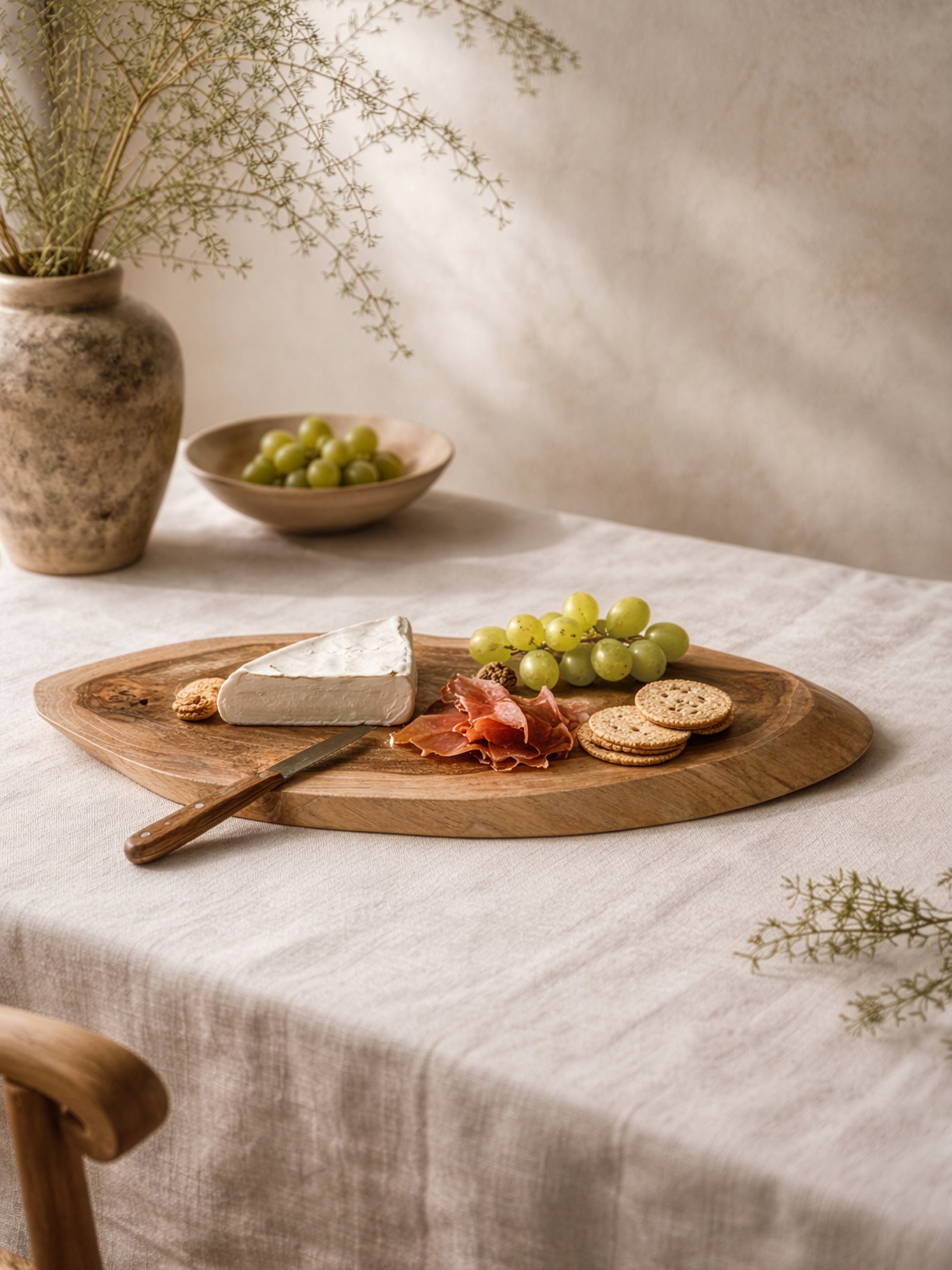 Vara cheese board in teak styled with cheese, grapes and crackers on a soft neutral table setting.