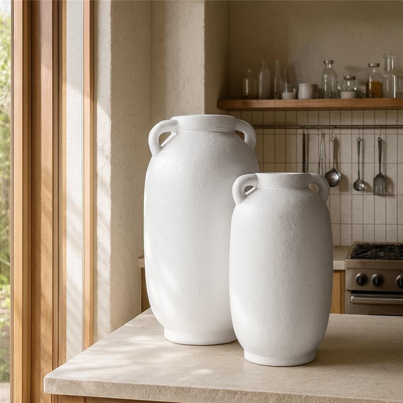 Pair of tall white amphora-style vases with twin handles, displayed on a stone kitchen bench in a light-filled, natural-toned kitchen.