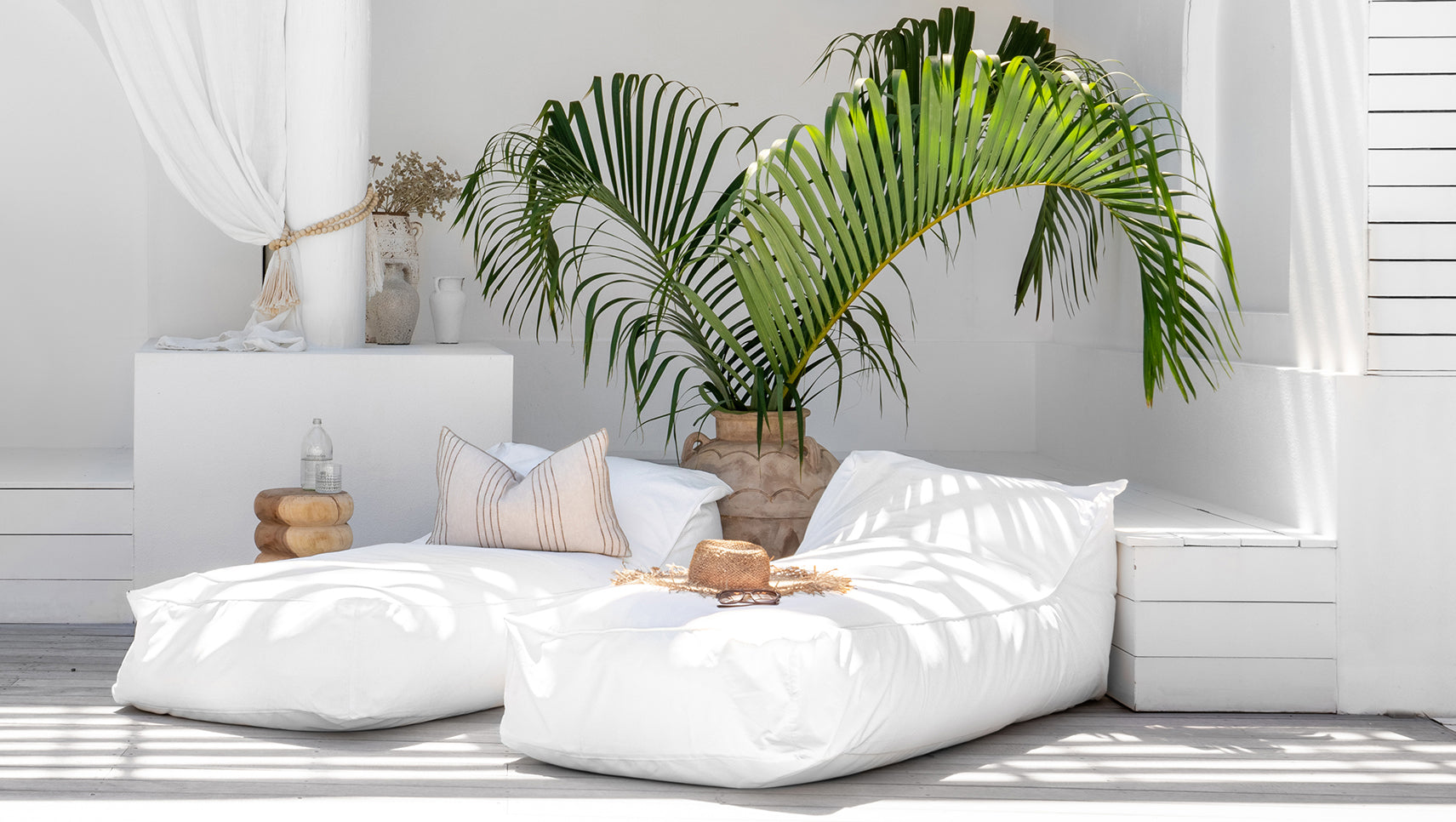 Two white bean bags styled with a striped cushion and straw hat, set in a tropical outdoor lounge area with lush palm leaves and white decor.