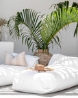 Two white bean bags styled with a striped cushion and straw hat, set in a tropical outdoor lounge area with lush palm leaves and white decor.