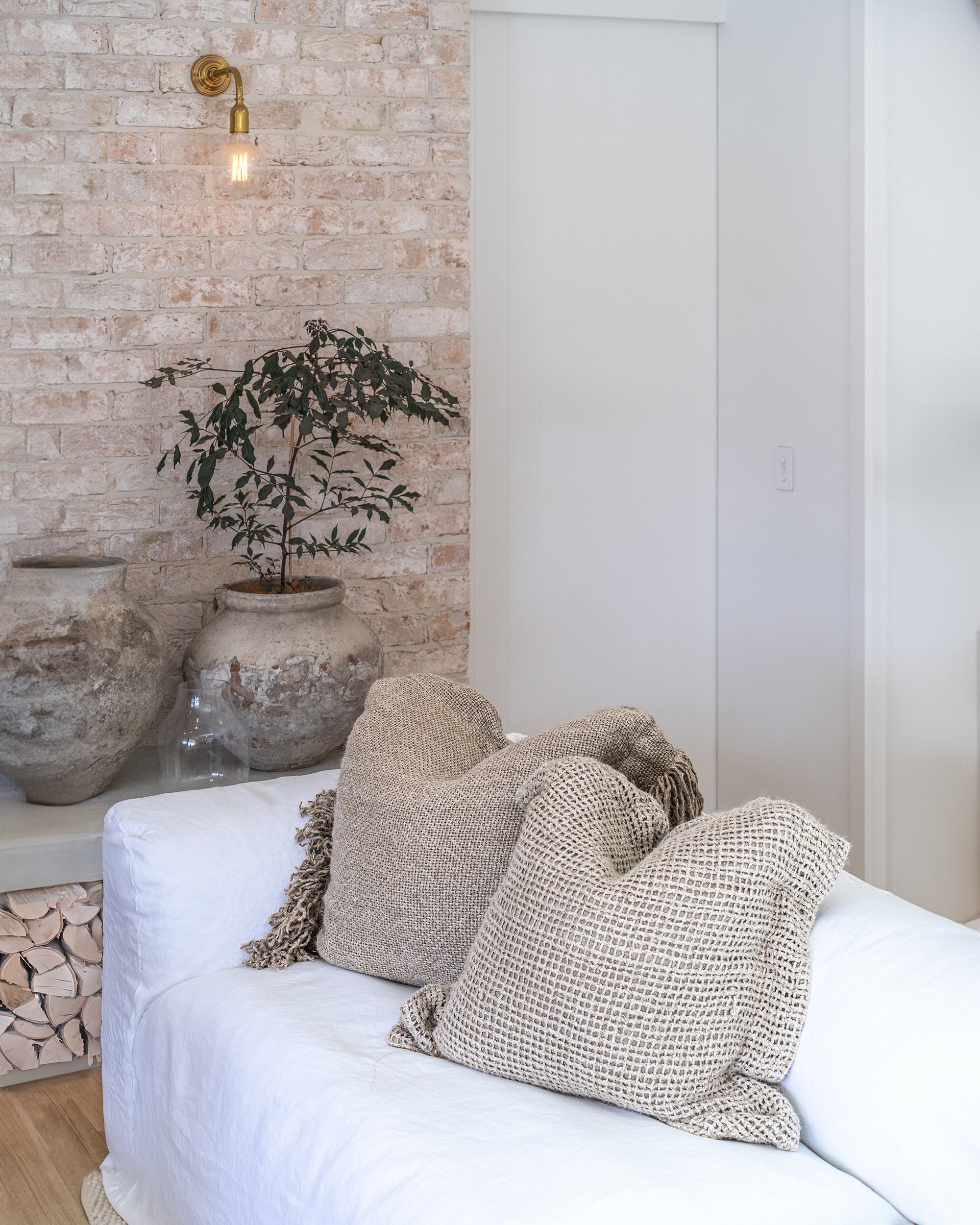 White linen sofa styled with textured neutral cushions, vintage ceramic pots, and stacked firewood against a rustic brick wall