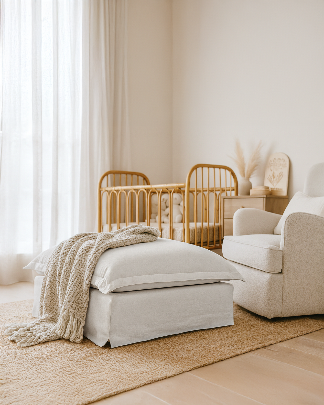 White linen ottoman with cushion and knitted throw in a neutral nursery, featuring a rattan cot, armchair, and warm natural decor.