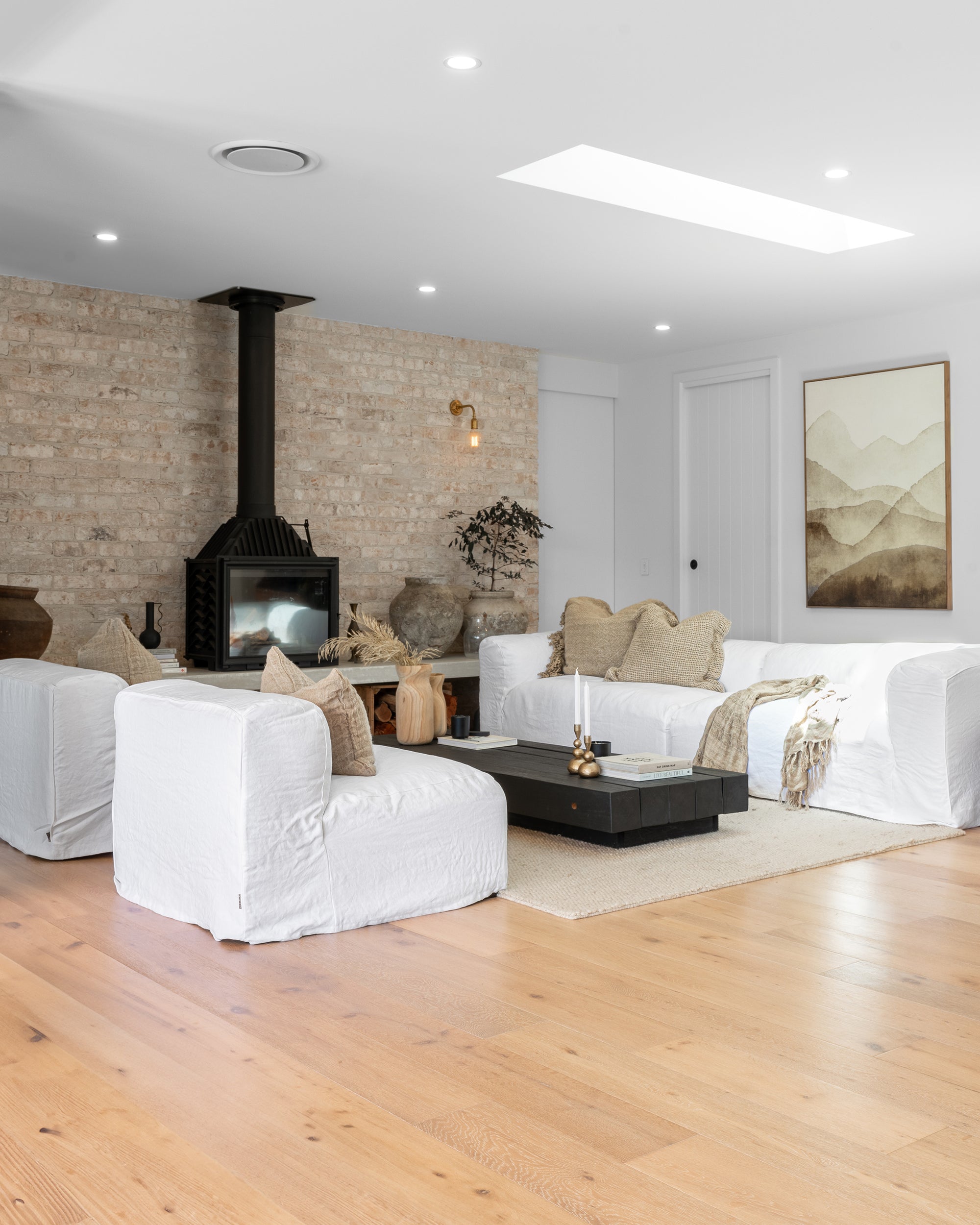 Bright and airy living room styled with white linen sofas, natural timber flooring, a black fireplace and soft neutral décor by KULALA Living.