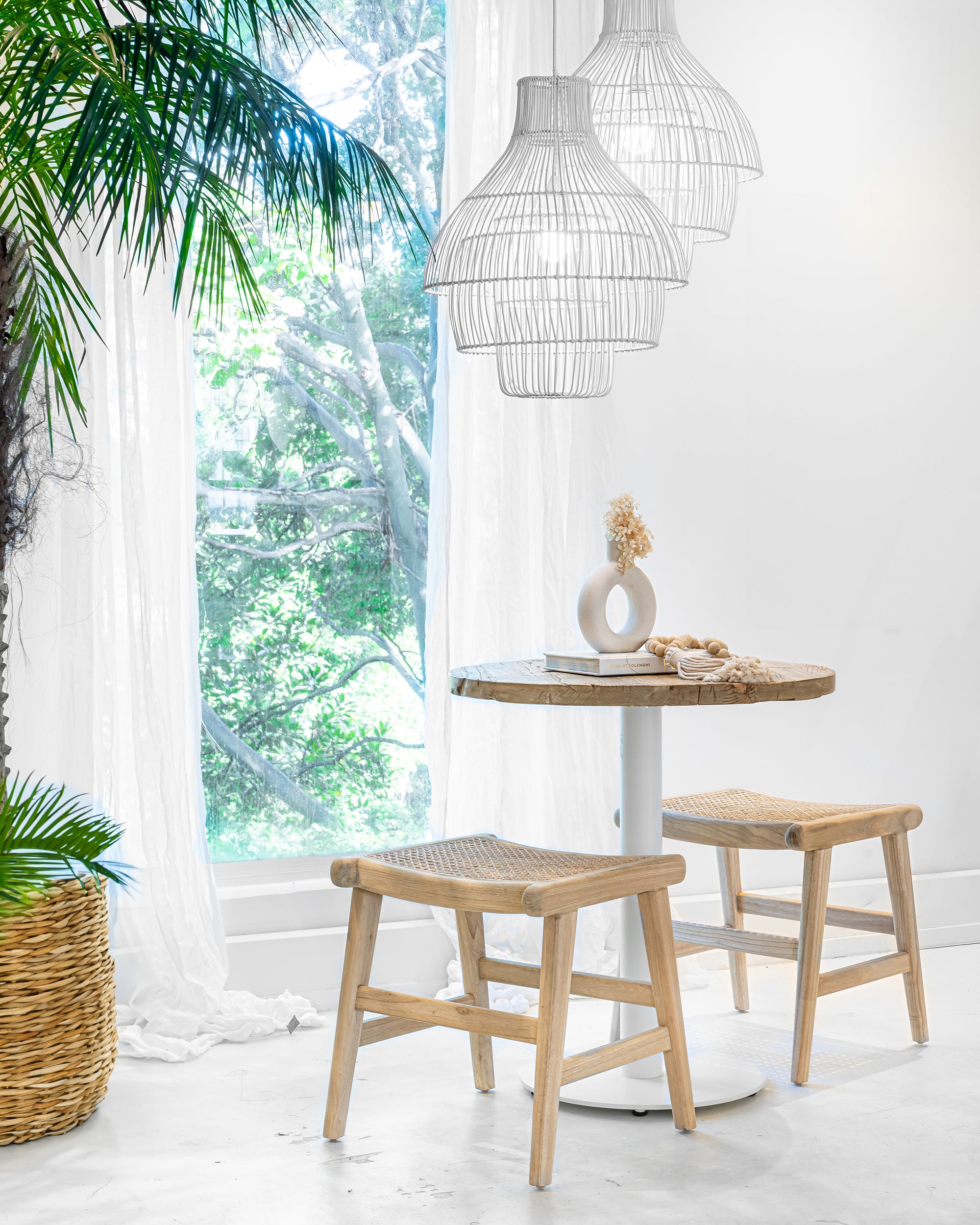 A serene dining nook featuring two white rattan pendant lights, a rustic round wooden table with a white pedestal base, and two natural wood stools with woven seats. Soft natural light filters through sheer curtains, enhancing the tranquil and airy aesthetic