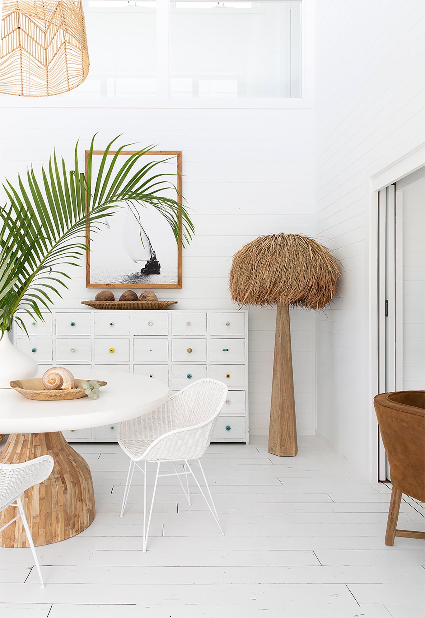 Bright coastal dining setting with white rattan dining chairs, natural wood round table, and woven pendant light. Styled with lush green plants and natural decor accents