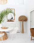 Bright coastal dining setting with white rattan dining chairs, natural wood round table, and woven pendant light. Styled with lush green plants and natural decor accents