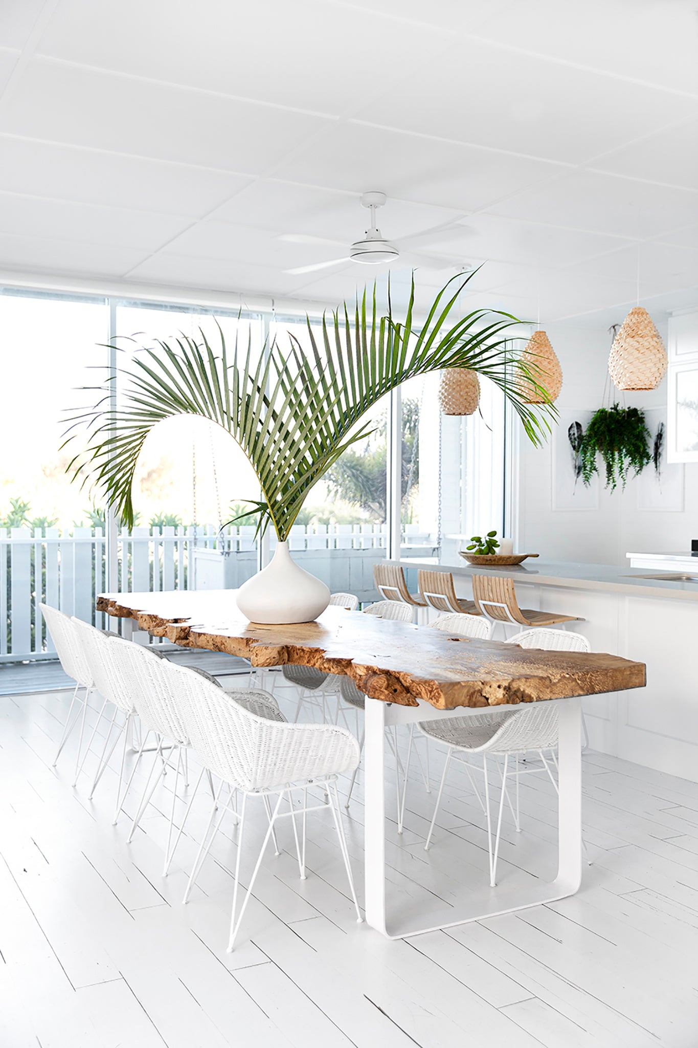 Bright coastal dining setting with white rattan dining chairs, natural wood round table, and woven pendant light. Styled with lush green plants and natural decor accents
