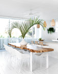 Bright coastal dining setting with white rattan dining chairs, natural wood round table, and woven pendant light. Styled with lush green plants and natural decor accents