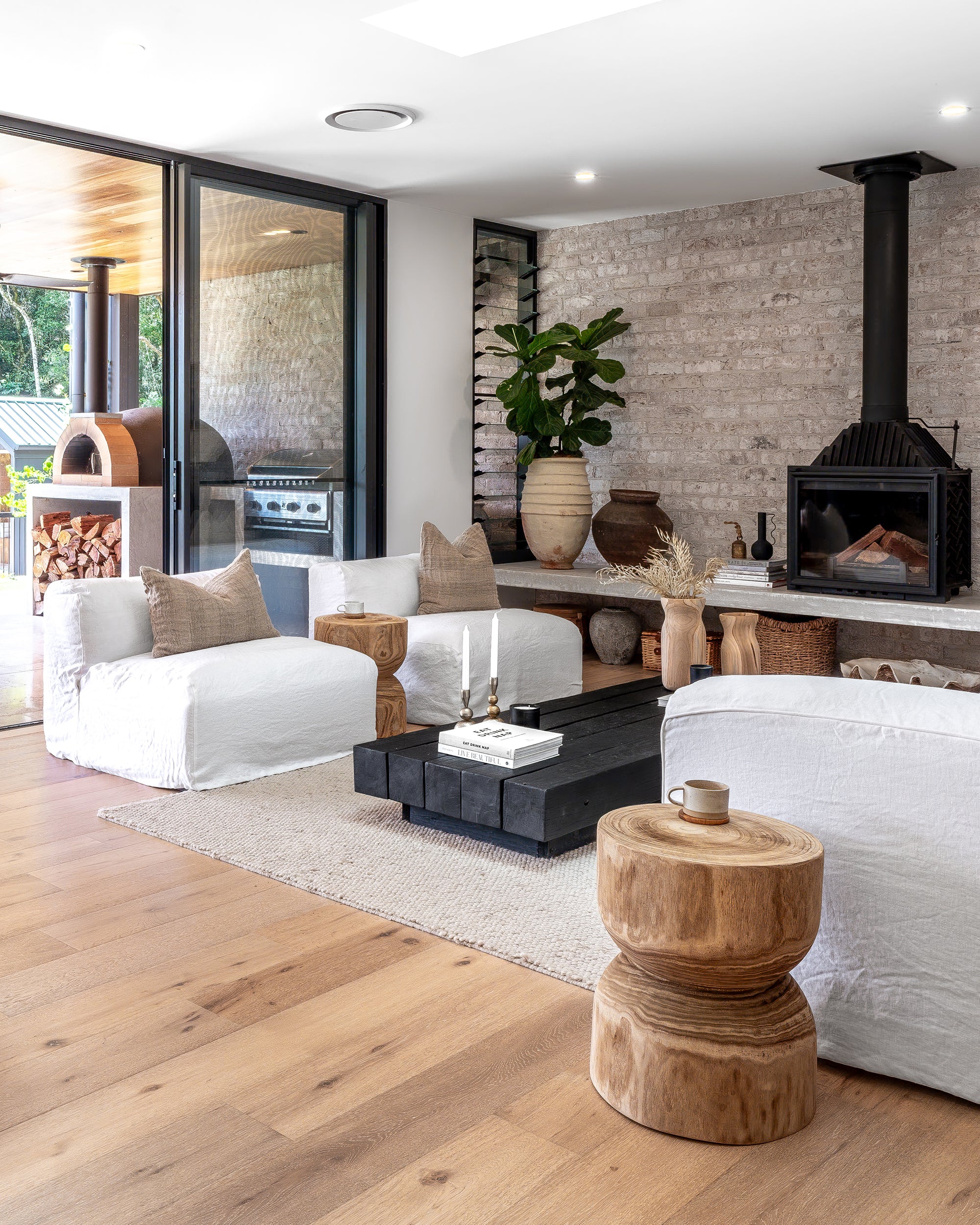 Light-filled living room featuring a white slipcover lounge, rustic stools, black timber coffee table, and a fireplace set against a natural brick wall, styled with earthy accents and neutral tones.