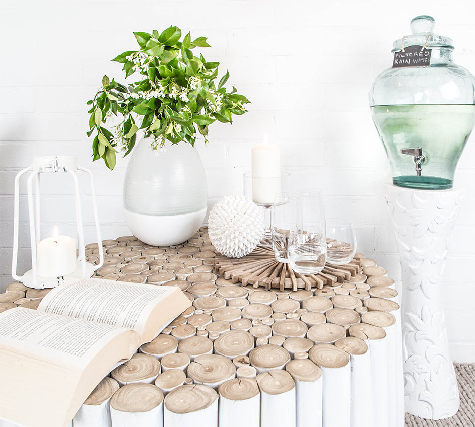 A beautifully styled setting featuring the Kulala Living White Teak Drum Coffee Table, adorned with an open book, candles, glassware, and a fresh greenery arrangement in a ceramic vase. The serene aesthetic is complemented by a glass water dispenser on a carved pedestal, creating a relaxed and inviting ambiance