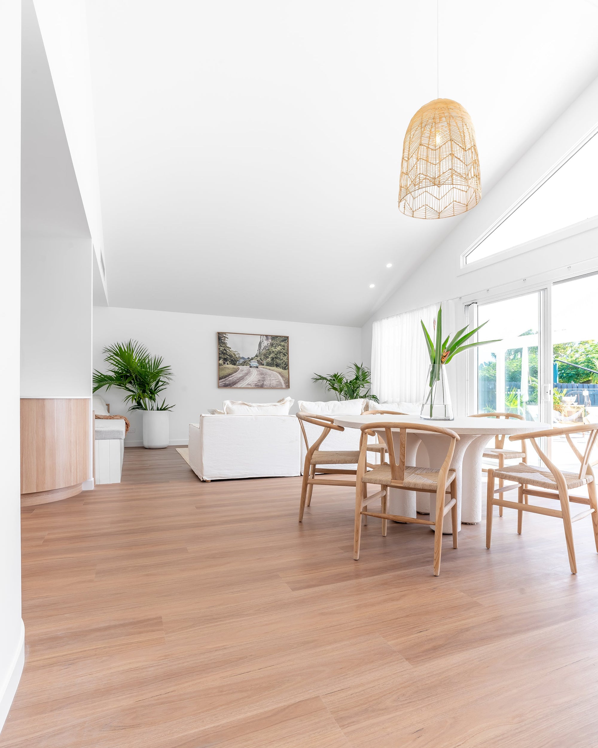 A bright and airy modern coastal dining space featuring natural timber wishbone dining chairs with woven seats, a curved white dining table, and soft neutral decor. Large windows invite natural light, complementing the organic textures and relaxed aesthetic