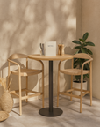 Natural teak wishbone-style bar chairs in a sunlit café-inspired setting with a round timber table and neutral-toned accessories