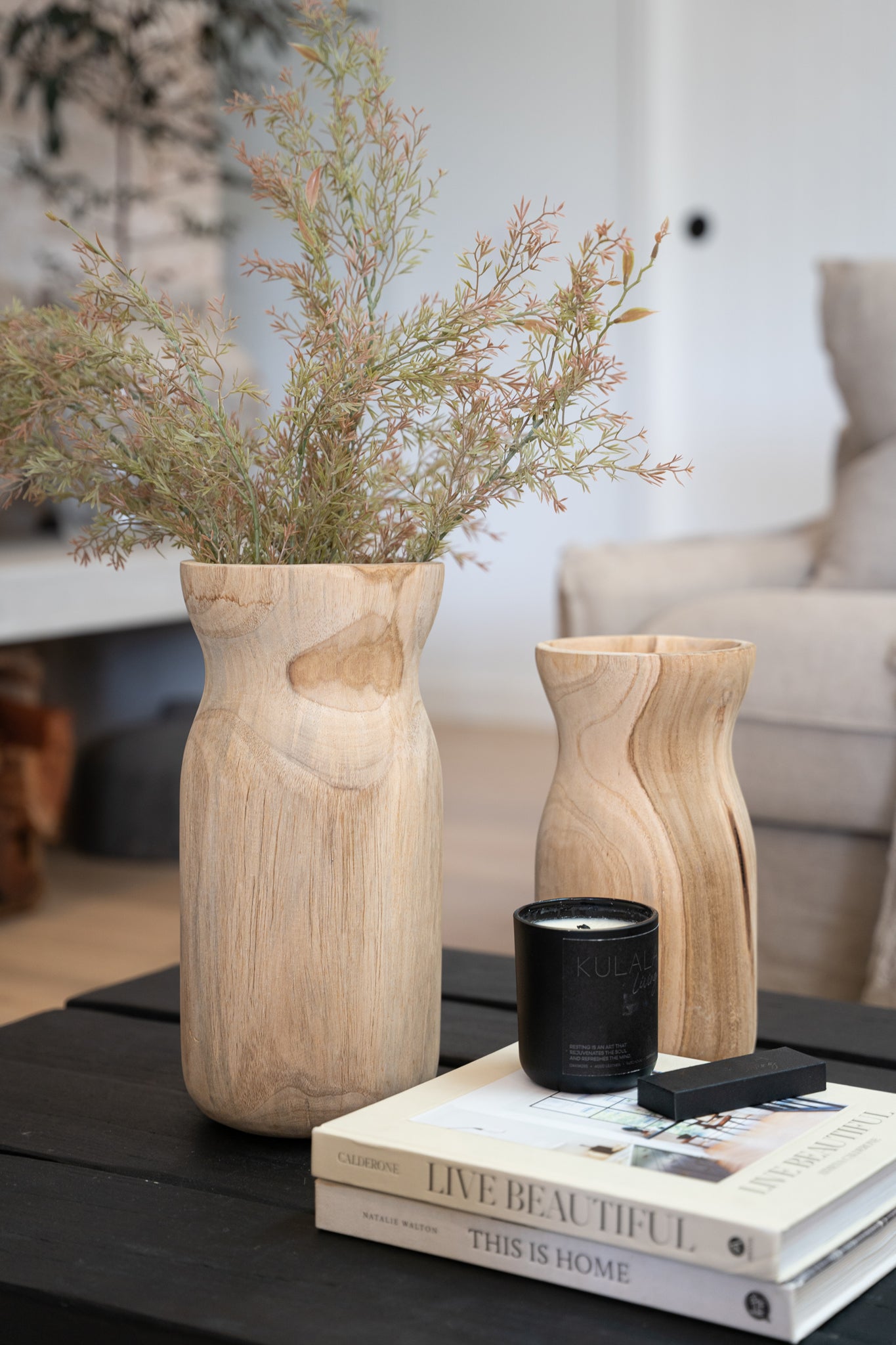 A beautifully styled coffee table featuring two sculptural wooden vases filled with delicate foliage, a black Kulala Living candle, and a curated stack of design books. The natural textures and earthy tones create a warm and inviting aesthetic, perfect for contemporary interiors