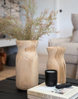 A beautifully styled coffee table featuring two sculptural wooden vases filled with delicate foliage, a black Kulala Living candle, and a curated stack of design books. The natural textures and earthy tones create a warm and inviting aesthetic, perfect for contemporary interiors